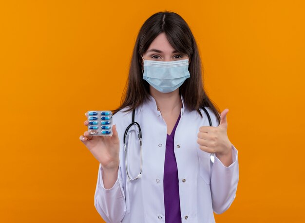Zelfverzekerde jonge vrouwelijke arts in medische mantel met stethoscoop draagt wegwerp medische gezichtsmasker houdt medicijnen en duimen omhoog op geïsoleerde oranje achtergrond met kopie ruimte