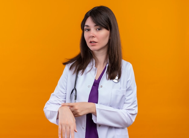 Zelfverzekerde jonge vrouwelijke arts in medische mantel met stethoscoop punten op tijd op geïsoleerde oranje achtergrond met kopie ruimte