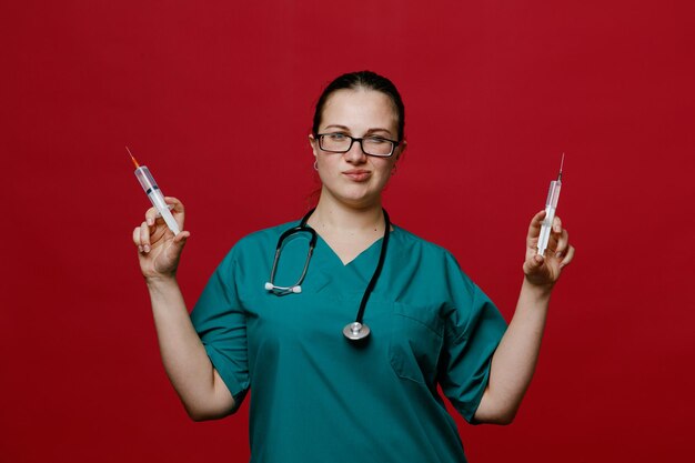 Zelfverzekerde jonge vrouwelijke arts met een uniforme bril en stethoscoop om haar nek met spuiten met naalden die naar camera kijken geïsoleerd op rode achtergrond