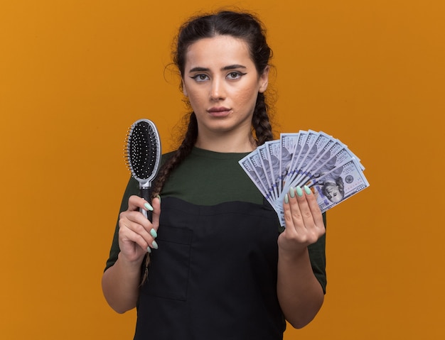 Gratis foto zelfverzekerde jonge vrouwelijke kapper in uniforme holdingskam met contant geld dat op oranje muur wordt geïsoleerd