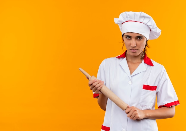 zelfverzekerde jonge vrouwelijke kok die de deegroller van de chef-kok uniforme holding op geïsoleerde gele muur met exemplaarruimte draagt