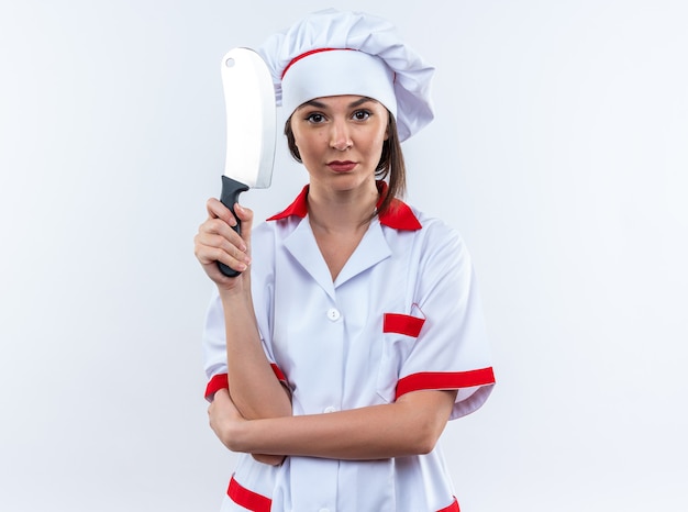Zelfverzekerde jonge vrouwelijke kok die een uniform van de chef-kok draagt met een hakmes geïsoleerd op een witte muur