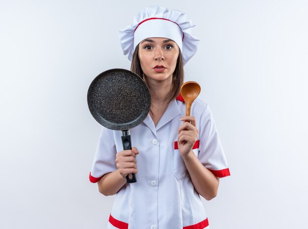 Zelfverzekerde jonge vrouwelijke kok die een uniform van de chef-kok draagt met een koekenpan met lepel geïsoleerd op een witte muur