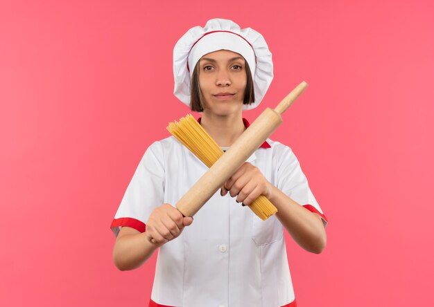 Zelfverzekerde jonge vrouwelijke kok in chef-kok uniform gebaren nee met spaghetti pasta en deegroller geïsoleerd op roze met kopie ruimte