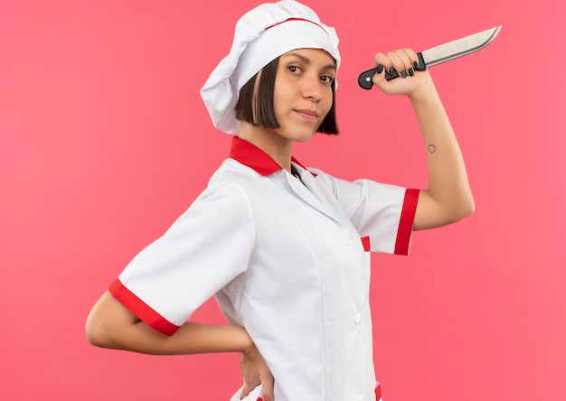 Zelfverzekerde jonge vrouwelijke kok in uniforme chef-kok staande in profiel te bekijken hand op taille zetten en mes geïsoleerd op roze te houden