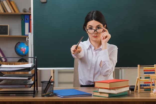 Zelfverzekerde jonge vrouwelijke leraar die een bril draagt, wijst naar de camera met de aanwijzer aan het bureau met schoolhulpmiddelen in de klas