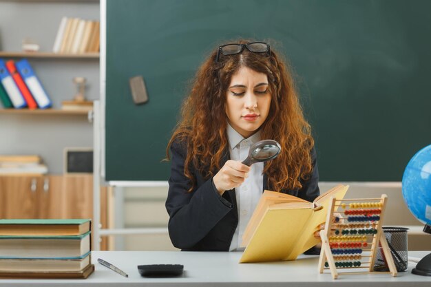 zelfverzekerde jonge vrouwelijke leraar met een bril die een boek leest met een vergrootglas dat aan het bureau zit met schoolhulpmiddelen in de klas