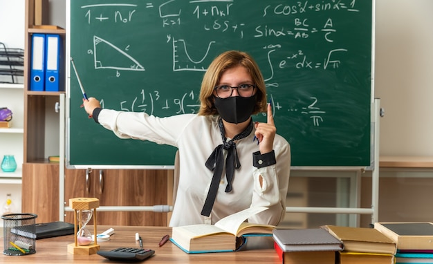 Zelfverzekerde jonge vrouwelijke leraar met een bril en een medisch masker zit aan tafel met schoolhulpmiddelen op het bord met de aanwijzer in de klas