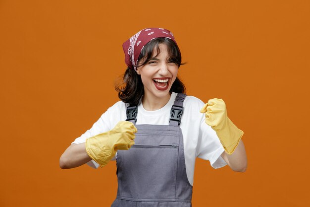Zelfverzekerde jonge vrouwelijke schoonmaker met uniforme rubberen handschoenen en bandana die naar de camera kijkt en een boksgebaar toont dat op een oranje achtergrond wordt geïsoleerd