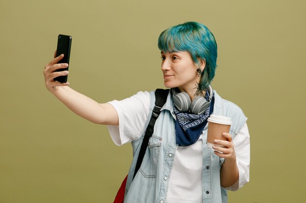 Zelfverzekerde jonge vrouwelijke student met koptelefoon en bandana op nek en rugzak met papieren koffiekopje selfie te nemen met mobiele telefoon geïsoleerd op olijfgroene achtergrond