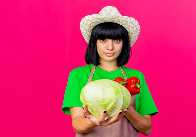 Zelfverzekerde jonge vrouwelijke tuinman in uniform dragen tuinieren hoed houdt kool en rode paprika