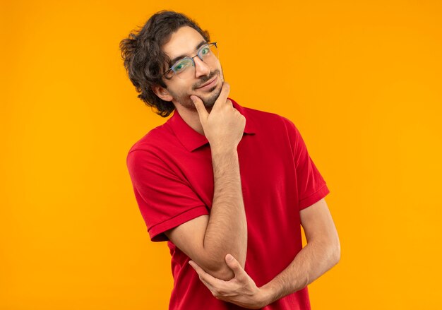 Zelfverzekerde jongeman in rood shirt met optische bril legt hand op kin geïsoleerd op oranje muur