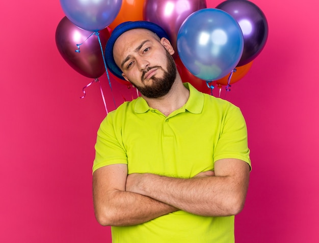 Gratis foto zelfverzekerde, kantelende jonge man met een blauwe feestmuts die voor ballonnen staat en handen kruist geïsoleerd op roze muur