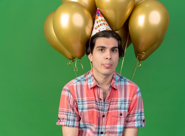 Zelfverzekerde knappe blanke man met verjaardagspet steekt tong uit en staat voor heliumballonnen