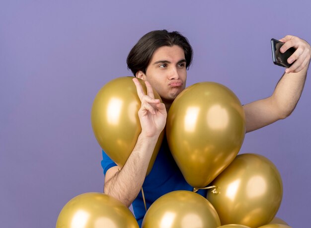 Zelfverzekerde knappe blanke man staat met heliumballonnen die een overwinningshandteken gebaren en naar de telefoon kijken die selfie maakt