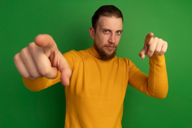 Gratis foto zelfverzekerde knappe blonde man wijst op camera met twee handen op groen