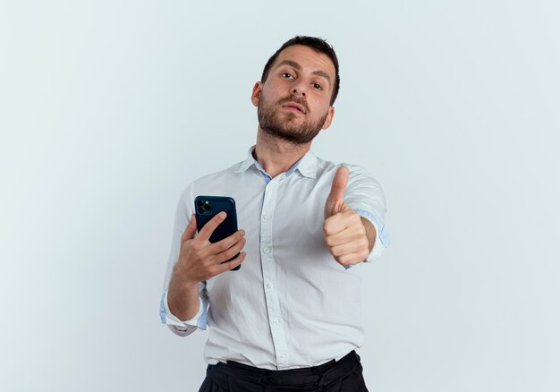Zelfverzekerde knappe man houdt telefoon en duimen omhoog geïsoleerd op een witte muur