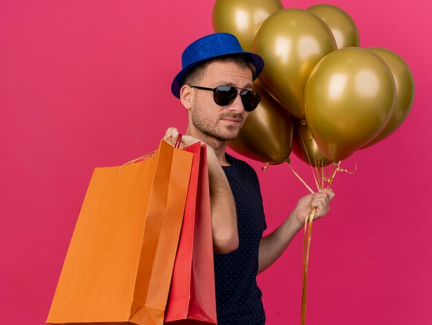 Zelfverzekerde knappe man in zonnebril met blauwe feestmuts staat zijwaarts met helium ballonnen en papieren boodschappentassen geïsoleerd op roze muur