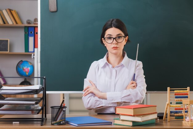 zelfverzekerde kruisende handen jonge vrouwelijke leraar die een bril draagt met de aanwijzer aan het bureau met schoolhulpmiddelen in de klas