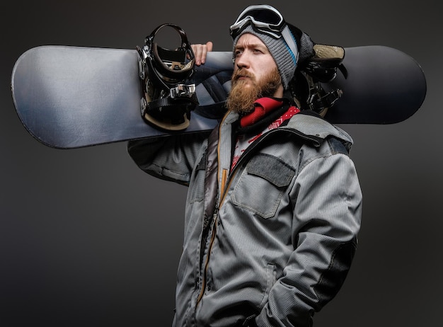 Gratis foto zelfverzekerde man met een rode baard met een volledige uitrusting die een snowboard op zijn schouder houdt, wegkijkend met een serieuze blik, geïsoleerd op een donkere achtergrond.