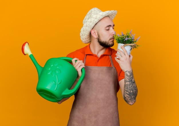 Zelfverzekerde mannelijke tuinman tuinieren hoed draagt houdt gieter en ruikt bloemen in bloempot