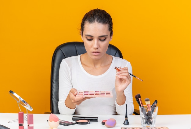 Zelfverzekerde, mooie blanke vrouw die aan tafel zit met make-uptools die een make-upborstel vasthoudt en naar een oogschaduwpalet kijkt