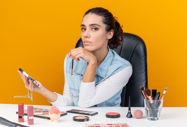 Zelfverzekerde mooie blanke vrouw zittend aan tafel met make-uptools met make-upborstel en spiegel geïsoleerd op een oranje muur met kopieerruimte