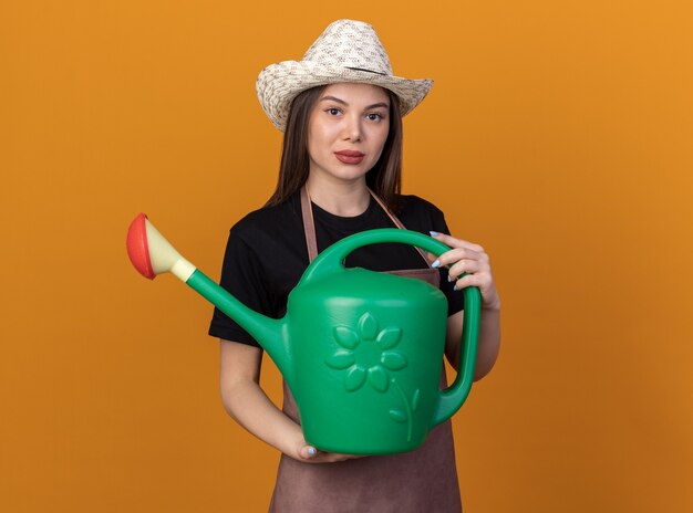 Zelfverzekerde, mooie blanke vrouwelijke tuinman met een tuinhoed met een gieter geïsoleerd op een oranje muur met kopieerruimte