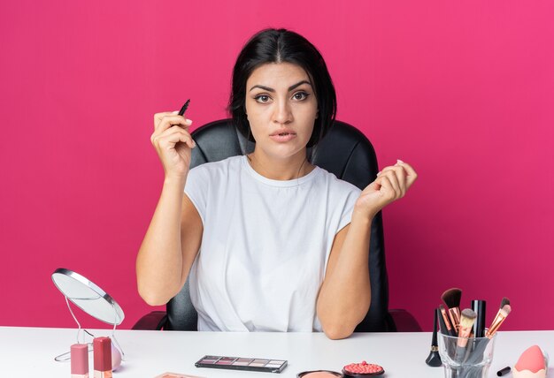 Zelfverzekerde mooie vrouw zit aan tafel met make-up tools met eyeliner