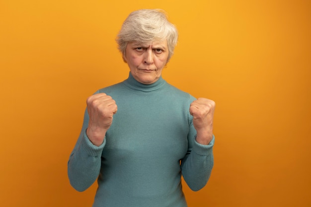 Gratis foto zelfverzekerde oude vrouw die blauwe coltrui draagt en naar voren balde vuisten doet, is een sterk gebaar geïsoleerd op een oranje muur met kopieerruimte