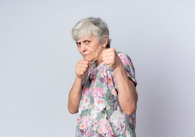 Zelfverzekerde oudere vrouw houdt vuisten klaar om te slaan geïsoleerd op een witte muur