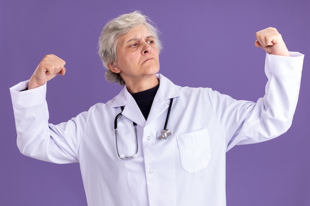 Zelfverzekerde oudere vrouw in doktersuniform met stethoscoop spant biceps aan en kijkt naar de zijkant