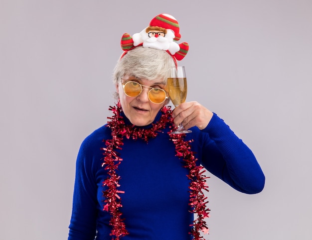 Zelfverzekerde oudere vrouw in zonnebril met santa hoofdband en slinger rond de nek met glas champagne geïsoleerd op een witte achtergrond met kopie ruimte