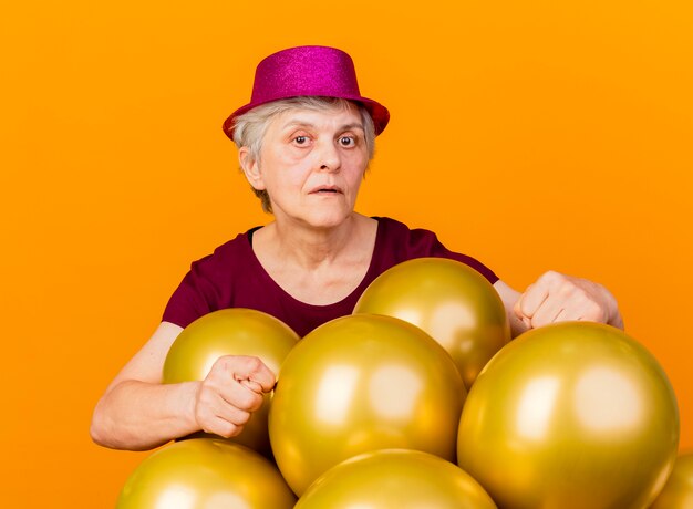Zelfverzekerde oudere vrouw met feestmuts staat met helium ballonnen gebaren badkamer handteken geïsoleerd op een oranje muur