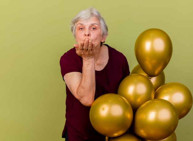 Zelfverzekerde oudere vrouw staat met helium ballonnen verzenden geïsoleerd op olijfgroene muur met kopie ruimte