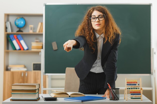 zelfverzekerde punten aan de zijkant jonge vrouwelijke leraar die voor het schoolbord staat in de klas