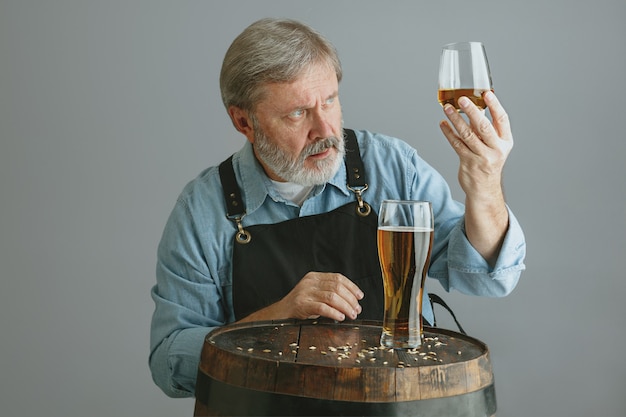 Gratis foto zelfverzekerde senior man brouwer met zelfgemaakt bier in glas op houten vat op grijs