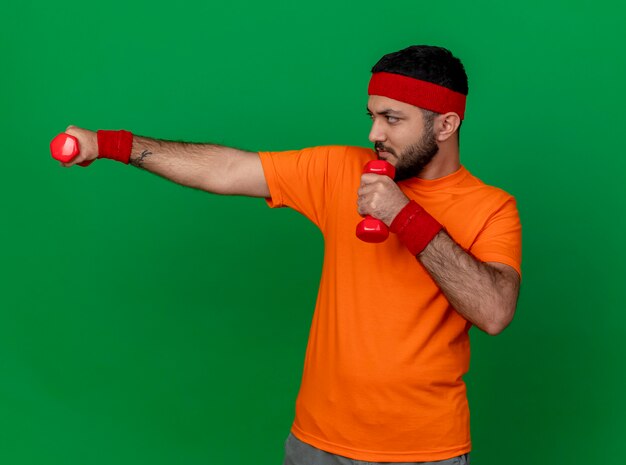 Zelfverzekerde sportieve jongeman in profiel te bekijken met hoofdband en polsband trainen met halters geïsoleerd op groene achtergrond