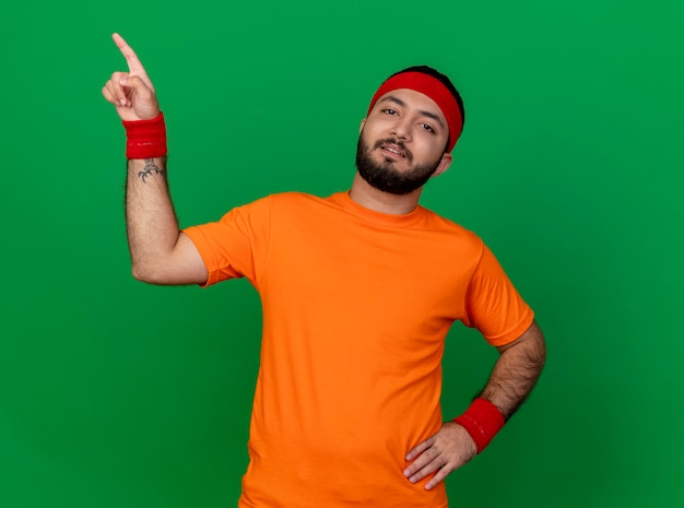 Gratis foto zelfverzekerde sportieve jongeman met hoofdband en polsband hand op heup en punten aan kant zetten geïsoleerd op groene achtergrond met kopie ruimte