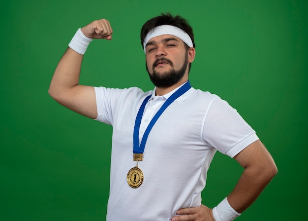 Zelfverzekerde sportieve jongeman met hoofdband en polsbandje met medaille met sterk gebaar geïsoleerd op groen