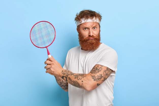 Zelfverzekerde sportman kijkt serieus, houdt tennisracket vast, heeft tatoeage op armen