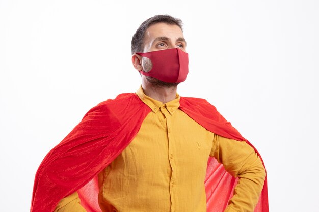 Zelfverzekerde superheld man met rode mantel dragen rood masker legt handen op taille en kijkt naar kant geïsoleerd op een witte muur