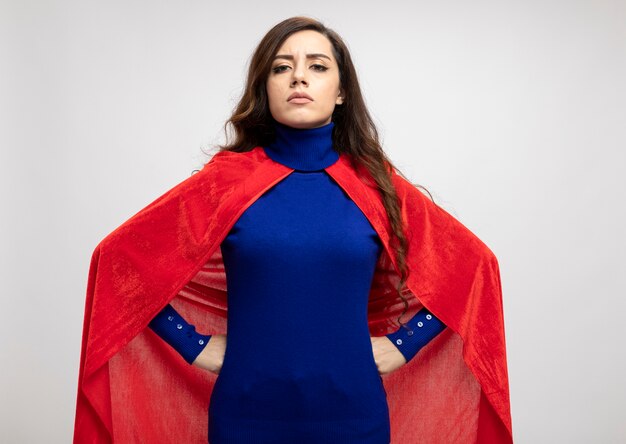 Zelfverzekerde supervrouw met rode cape legt handen op taille geïsoleerd op een witte muur