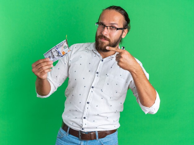 Zelfverzekerde volwassen knappe man met een bril die vasthoudt en wijst op een brandende dollar die naar een camera kijkt die op een groene muur is geïsoleerd