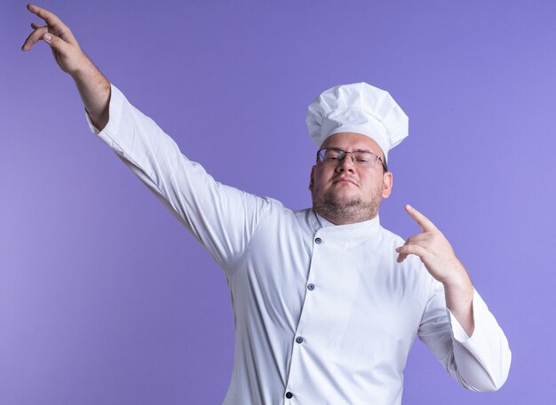 Zelfverzekerde volwassen mannelijke kok met een uniform van de chef-kok en een bril die naar de voorkant kijkt en omhoog wijst op een paarse muur