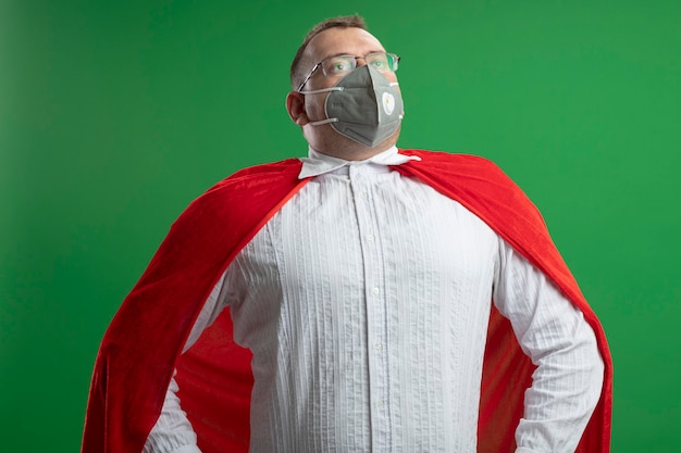 Gratis foto zelfverzekerde volwassen slavische superheld man in rode cape bril en beschermend masker houden handen op taille opzoeken geïsoleerd op groene muur