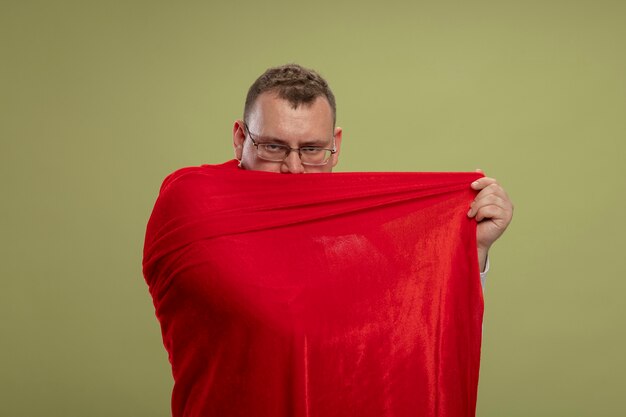 Zelfverzekerde volwassen Slavische superheld man in rode cape die een bril draagt die zichzelf bedekt met cape van erachter geïsoleerd op olijfgroene muur