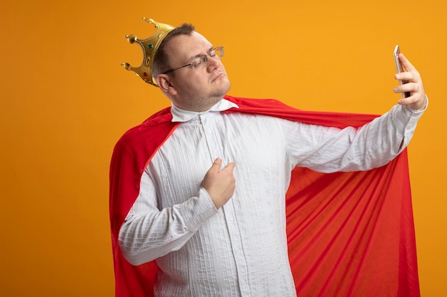 Gratis foto zelfverzekerde volwassen superheld man in rode cape bril en kroon wijzend op zichzelf nemen selfie geïsoleerd op oranje muur