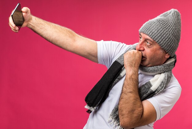 Zelfverzekerde volwassen zieke blanke man met sjaal om nek met wintermuts kijkend naar telefoon selfie nemend en vuist dicht bij mond houden geïsoleerd op roze muur met kopieerruimte