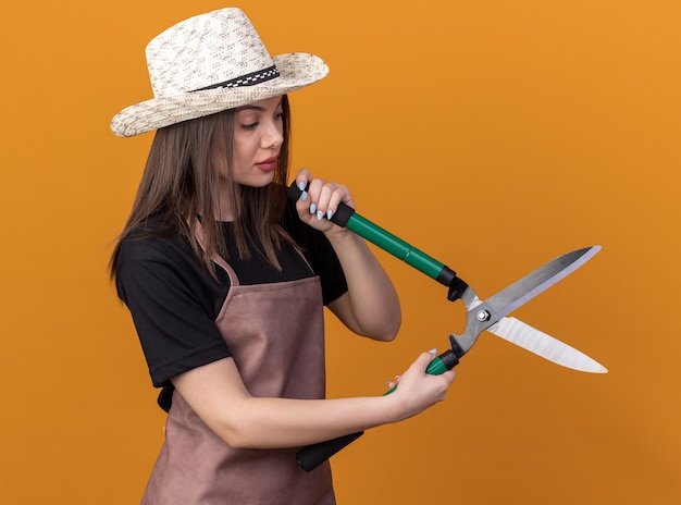 Gratis foto zelfverzekerde, vrij blanke vrouwelijke tuinman die een tuinhoed draagt en naar een tuinschaar kijkt